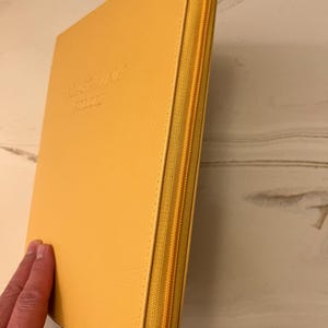 以下が含まれることがあります： 前面に「MAGAZINE JOURNAL」という文字がエンボス加工された、黄色の革製のジッパー付きバインダーです。