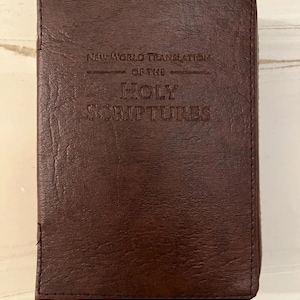 以下が含まれることがあります： 「New World Translation of the Holy Scriptures」のエンボス加工が施された茶色の革装丁の本。本は長方形で、閉じられているように見えます。表紙は質感のある表面です。