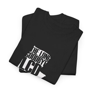 Op de afbeelding: Twee zwarte t-shirts zijn netjes opgevouwen en op elkaar gestapeld. Het bovenste shirt heeft de witte tekst "THE LONG GOODBYE LCD" op de voorkant. De shirts zijn gemaakt van een zacht materiaal, waarschijnlijk katoen of een katoenmix.