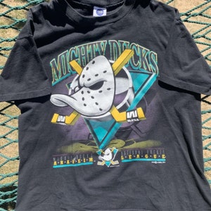 Op de afbeelding: Zwart T-shirt met een afbeelding van een cartoon eend die een hockeymasker draagt en hockeysticks vasthoudt. De tekst "Mighty Ducks Anaheim" en "National Hockey League" staat op het shirt gedrukt.