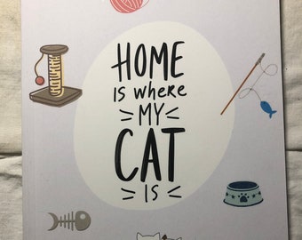Puede incluir: Una libreta blanca y gris claro con el texto "Home is where my cat is" en letras negras. La libreta presenta ilustraciones de un gato, un juguete para gatos, un cuenco de comida, una espina de pescado y una bola de lana.