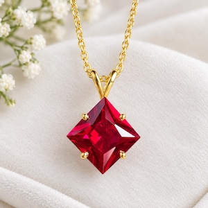 May include: A gold chain necklace featuring a square-cut, deep red gemstone pendant held by four prongs. The pendant is oriented in a diamond shape, showcasing a faceted surface that reflects light, suspended from a delicate gold link chain.