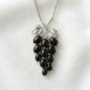 May include: Silver-toned necklace featuring a pendant shaped like a bunch of grapes. The pendant has a silver vine with two leaves and a cluster of black, oval-shaped beads. The necklace hangs against a white background.