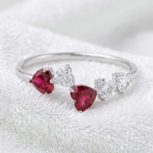 May include: A silver ring with heart-shaped gemstones. The ring showcases two vivid red rubies and three clear diamonds. The gemstones are arranged in a delicate, alternating pattern, creating a romantic and elegant design.