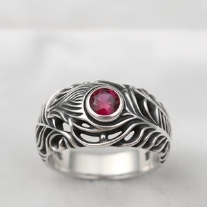 May include: A silver ring with an ornate feather design. The ring features a round, red gemstone at its center. The band has a detailed, openwork pattern. The ring is a piece of jewelry.