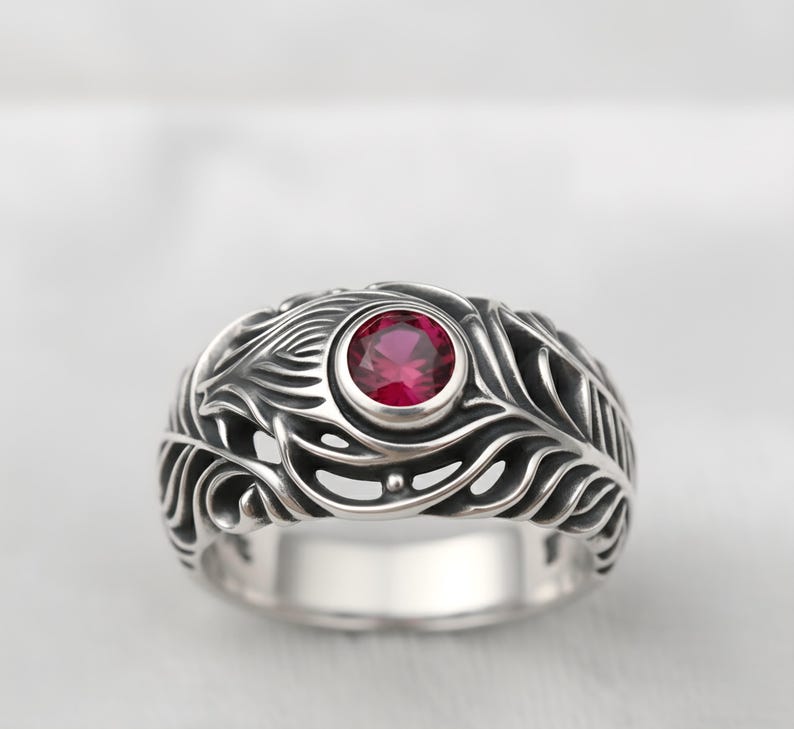 May include: A silver ring with an ornate feather design. The ring features a round, red gemstone at its center. The band has a detailed, openwork pattern. The ring is a piece of jewelry.