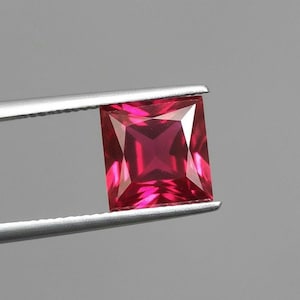 May include: A square-cut, vibrant red gemstone held by silver tweezers. The gemstone has a faceted surface that reflects light, showcasing its internal structure and color. The gem is a deep, rich shade of red.