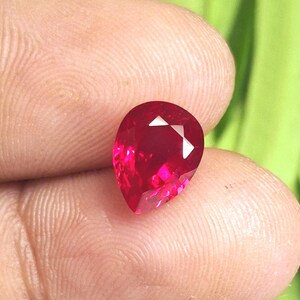 Puede incluir: Una gema de rubí facetada en forma de pera. La gema es de color rojo vibrante con una superficie pulida, que muestra reflejos internos. La gema se sostiene entre los dedos, con un fondo verde visible.