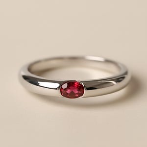 Può includere: Un anello in argento lucido con una vibrante pietra preziosa di rubino taglio ovale. L'anello ha una fascia liscia e arrotondata e un'incastonatura a castone che tiene saldamente la gemma rossa. Il design è semplice ed elegante.