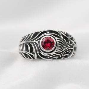 May include: A silver ring with a central, round, red gemstone. The ring features an intricate feather-like design around the gemstone. The ring is set against a white background, highlighting the details of the jewelry.