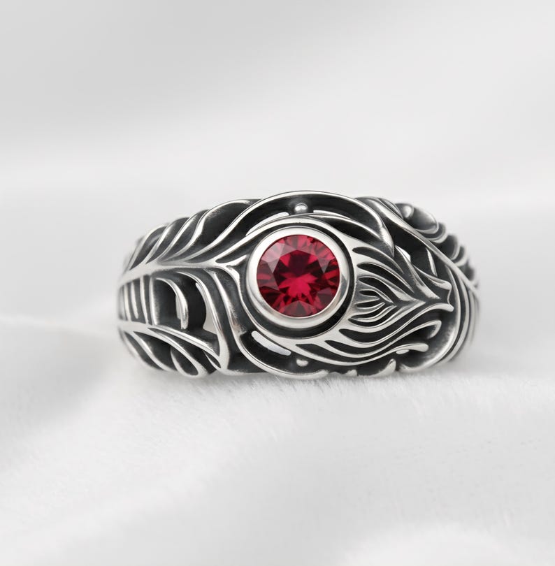 May include: A silver ring with a central, round, red gemstone. The ring features an intricate feather-like design around the gemstone. The ring is set against a white background, highlighting the details of the jewelry.