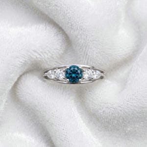 May include: A silver-toned ring featuring a central round-cut dark blue gemstone flanked by two smaller round-cut clear stones and two outer pear-shaped clear stones, all set within a polished metal band with a smooth, rounded profile.