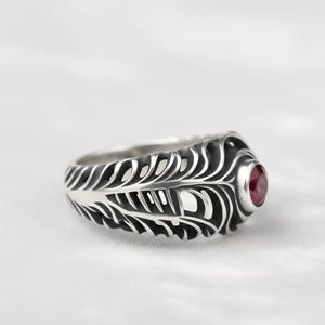 May include: A silver ring with a feather design and a round, red gemstone. The ring has intricate detailing, with the feather pattern extending around the band. The gemstone is set in a bezel setting, adding a pop of color to the ring.