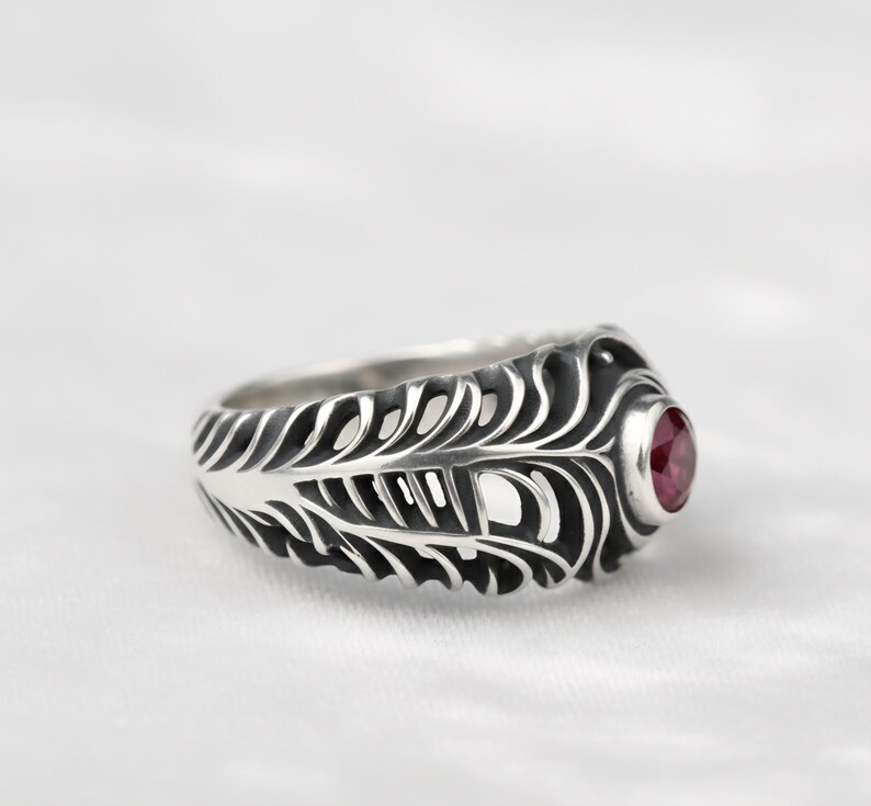 May include: A silver ring with a feather design and a round, red gemstone. The ring has intricate detailing, with the feather pattern extending around the band. The gemstone is set in a bezel setting, adding a pop of color to the ring.