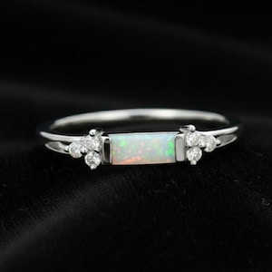 May include: A silver ring featuring a rectangular opal centre stone and small, round accent stones. The opal has a white base with green and orange hues. The ring is set against a black background.