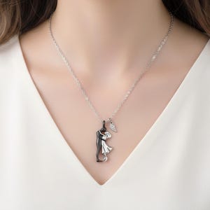 K&ouml;nnte beinhalten: Eine silberne Halskette mit einem Anh&auml;nger, der ein sich umarmendes Paar zeigt, eine Figur in Schwarz und die andere in Wei&szlig;. Ein kleiner Fl&uuml;gelanh&auml;nger h&auml;ngt neben dem Paar. Die Halskette wird &uuml;ber einem wei&szlig;en Oberteil getragen.