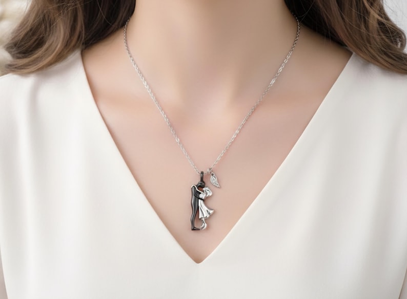 K&ouml;nnte beinhalten: Eine silberne Halskette mit einem Anh&auml;nger, der ein sich umarmendes Paar zeigt, eine Figur in Schwarz und die andere in Wei&szlig;. Ein kleiner Fl&uuml;gelanh&auml;nger h&auml;ngt neben dem Paar. Die Halskette wird &uuml;ber einem wei&szlig;en Oberteil getragen.