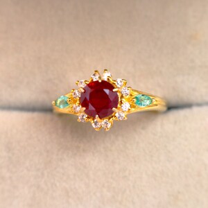 Peut inclure: Une bague en or ornée d'une grosse pierre rouge foncé et ronde au centre, entourée de petites pierres blanches. Deux pierres vertes en forme de goutte d'eau flanquent la pierre centrale. La bague a un design détaillé et orné.