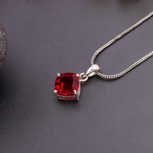 Pode incluir: Um colar de prata com um pingente quadrado de pedra preciosa vermelho escuro. O pingente é fixado em uma moldura de prata e pendurado em uma corrente de prata fina. A pedra preciosa tem uma superfície polida e o design é simples e elegante.