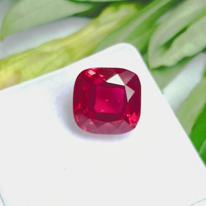 May include: A faceted, cushion-cut ruby gemstone. The ruby is a deep, vibrant red colour and sits on a white surface. Green leaves are visible in the background.