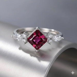 Può includere: Un anello d'argento con una gemma rosso scuro quadrata al centro. L'anello è impreziosito da gemme più piccole e trasparenti e da pietre a forma di pera su entrambi i lati della pietra principale. L'anello è montato su una fascia d'argento.