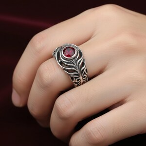 May include: A silver ring with a red gemstone set in a decorative, openwork design. The ring features a central, round red stone, surrounded by a detailed, feather-like pattern. The ring is on a finger.