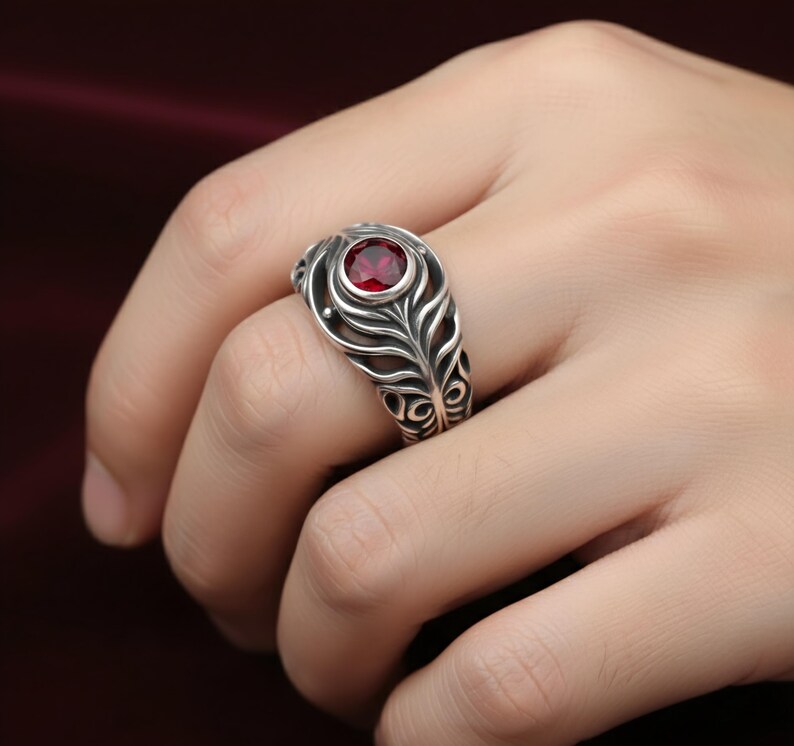 May include: A silver ring with a red gemstone set in a decorative, openwork design. The ring features a central, round red stone, surrounded by a detailed, feather-like pattern. The ring is on a finger.