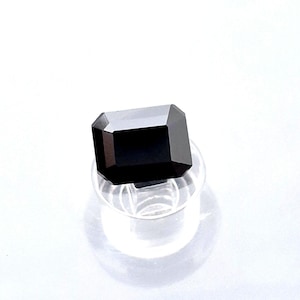 May include: A faceted, rectangular black gemstone, possibly an onyx or similar stone, is displayed on a clear, circular stand. The gem's polished surfaces reflect light, showcasing its geometric cut and deep color. The background is a bright white.