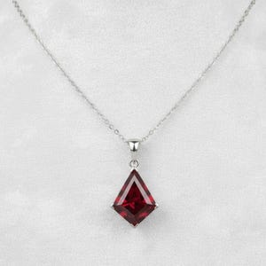 Op de afbeelding: Een zilveren ketting met een diamantvormige, dieprode edelsteen hanger. De hanger is facetgeslepen en gezet in een zilveren montuur, hangend aan een fijne zilveren ketting. De achtergrond is effen wit.