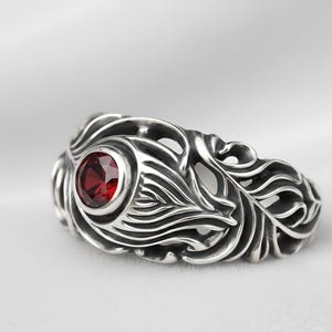 May include: A silver ring with an ornate, flowing design. The ring features a round, deep red gemstone set in the center. The metalwork has a detailed, textured pattern, creating a unique and eye-catching piece of jewelry.