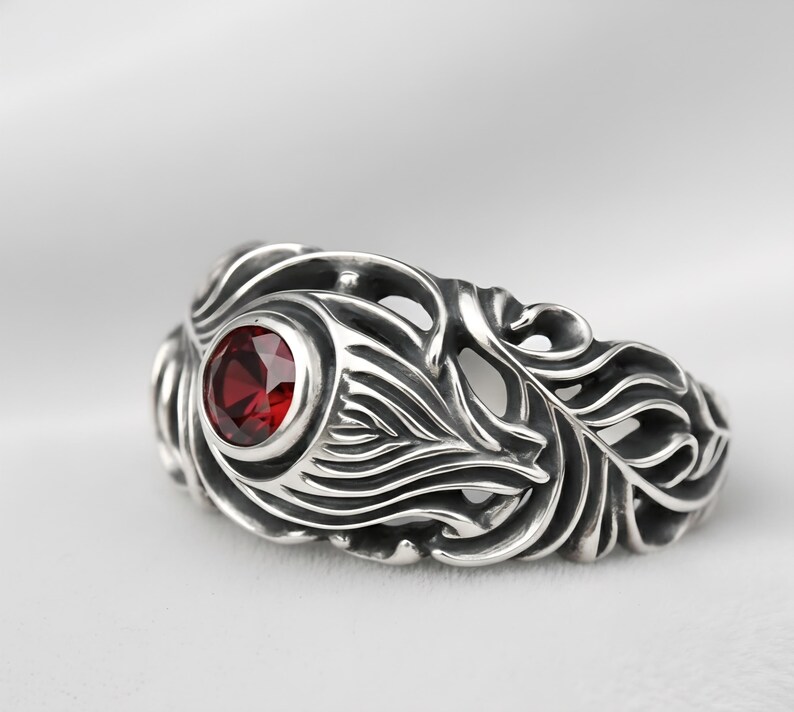 May include: A silver ring with an ornate, flowing design. The ring features a round, deep red gemstone set in the center. The metalwork has a detailed, textured pattern, creating a unique and eye-catching piece of jewelry.