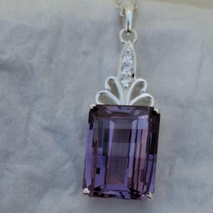 Peut inclure: Un collier pendentif en argent avec une grande pierre précieuse violette rectangulaire et facettée. Le pendentif a un dessus décoratif en argent avec trois petites pierres claires. Le collier est sur fond gris doux.