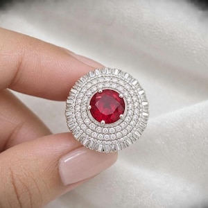 May include: A circular ring featuring a prominent round-cut red gemstone at the centre, surrounded by multiple concentric halos of small, clear, round-cut stones and an outer border of baguette-cut stones set in a polished silver-toned metal band.