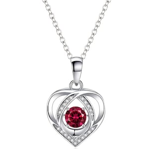 Può includere: Una collana d'argento con un ciondolo a forma di cuore. Il ciondolo presenta una grande gemma rotonda rosso rubino al centro, circondata da un alone di piccoli cristalli trasparenti. La catena della collana è delicata e argentata.