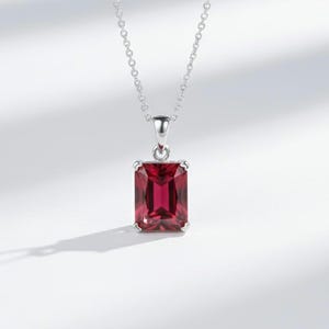 May include: A silver necklace with a rectangular, deep red gemstone pendant. The pendant is set in a silver frame and hangs from a delicate chain. The gemstone has a faceted surface, reflecting light.
