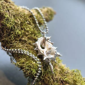 May include: A silver-toned pendant shaped like a spiky, organic shell with a hollow center, suspended from a metallic ball chain. The pendant features a textured, metallic finish with multiple pointed protrusions extending from its central core.