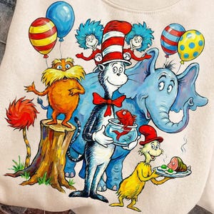 Puede incluir: Sudadera de color crema con una colorida ilustración de personajes de un libro infantil. El diseño incluye un elefante azul, un gato con sombrero rojo y blanco y otras figuras caprichosas, junto con globos.
