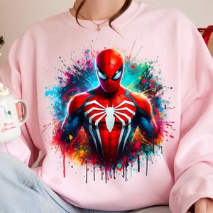 Puede incluir: Sudadera rosa con un estampado gr&aacute;fico colorido de Spiderman en un traje rojo y azul con un logotipo de ara&ntilde;a blanco. El gr&aacute;fico tiene un efecto de pintura salpicada.