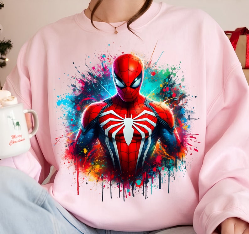 Puede incluir: Sudadera rosa con un estampado gr&aacute;fico colorido de Spiderman en un traje rojo y azul con un logotipo de ara&ntilde;a blanco. El gr&aacute;fico tiene un efecto de pintura salpicada.
