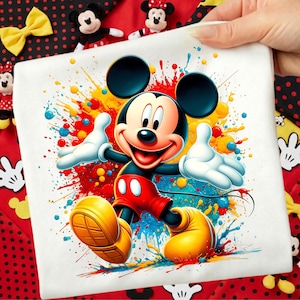 Peut inclure: Un carré blanc avec une illustration vibrante de Mickey Mouse sur un fond éclaboussé de peinture colorée. Mickey est représenté dans sa tenue classique : short rouge, chaussures jaunes et gants blancs. L'image est tenue par une main, avec des peluches Mickey et Minnie Mouse en arrière-plan.
