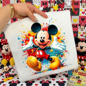K&ouml;nnte beinhalten: Wei&szlig;es quadratisches Kissen mit einem lebendigen Mickey Mouse-Design. Mickey ist in seiner klassischen Kleidung dargestellt, umgeben von bunten Farbspritzern. Das Kissen wird von einer Hand gehalten, mit anderen Mickey Mouse-Artikeln im Hintergrund.