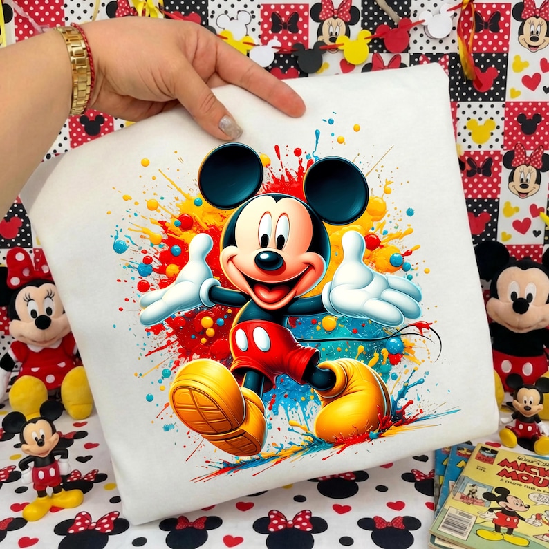 K&ouml;nnte beinhalten: Wei&szlig;es quadratisches Kissen mit einem lebendigen Mickey Mouse-Design. Mickey ist in seiner klassischen Kleidung dargestellt, umgeben von bunten Farbspritzern. Das Kissen wird von einer Hand gehalten, mit anderen Mickey Mouse-Artikeln im Hintergrund.