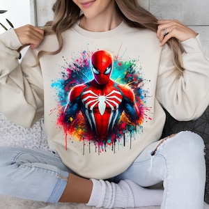Puede incluir: Una sudadera beige con un estampado gr&aacute;fico colorido de Spiderman. El estampado presenta a Spiderman con un traje rojo y azul con un logotipo de ara&ntilde;a negro. El fondo es una salpicadura de pintura colorida.