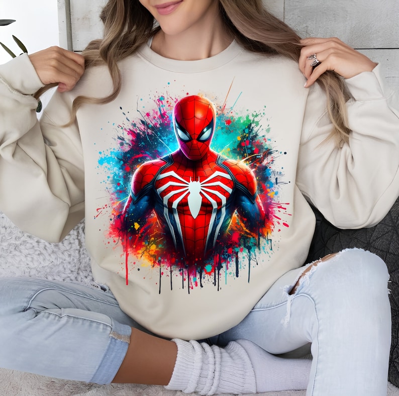 Puede incluir: Una sudadera beige con un estampado gr&aacute;fico colorido de Spiderman. El estampado presenta a Spiderman con un traje rojo y azul con un logotipo de ara&ntilde;a negro. El fondo es una salpicadura de pintura colorida.