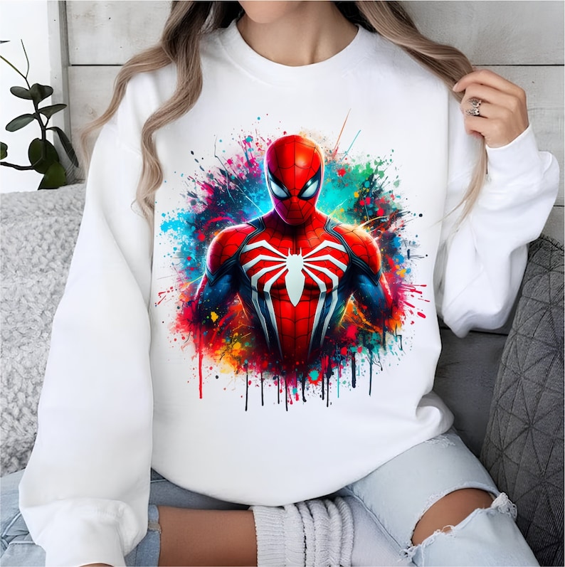 Puede incluir: Sudadera blanca con un estampado gr&aacute;fico colorido de Spiderman en un traje rojo y azul con un logotipo de ara&ntilde;a en el pecho. El gr&aacute;fico tiene un efecto de salpicadura de pintura.