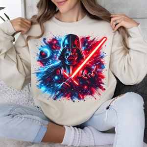 May include: A cream-colored sweatshirt with a graphic print of Darth Vader holding a red lightsaber. The background is a blue and red galaxy with stars and planets.