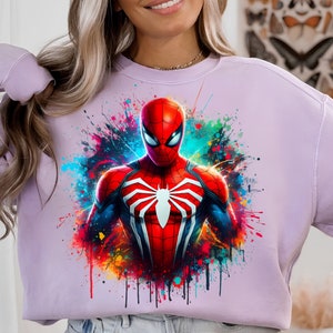 Puede incluir: Una sudadera de color lavanda con un estampado gr&aacute;fico colorido de Spider-Man con un traje rojo y negro. El gr&aacute;fico tiene un efecto de pintura salpicada.