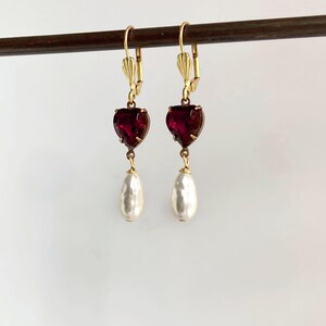 May include: A pair of gold-tone leverback earrings featuring a faceted deep red heart-shaped stone suspended above a white teardrop-shaped pearl-like bead. The earrings are displayed hanging from a thin horizontal dark brown rod.
