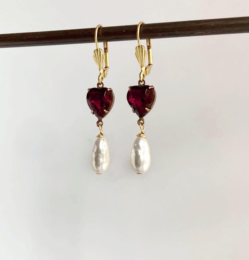 May include: A pair of gold-tone leverback earrings featuring a faceted deep red heart-shaped stone suspended above a white teardrop-shaped pearl-like bead. The earrings are displayed hanging from a thin horizontal dark brown rod.