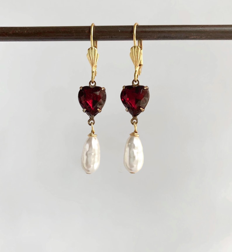 May include: A pair of gold-tone leverback earrings featuring a faceted deep red heart-shaped stone suspended above a white teardrop-shaped pearl. The earrings are displayed hanging from a thin horizontal rod against a plain light background.